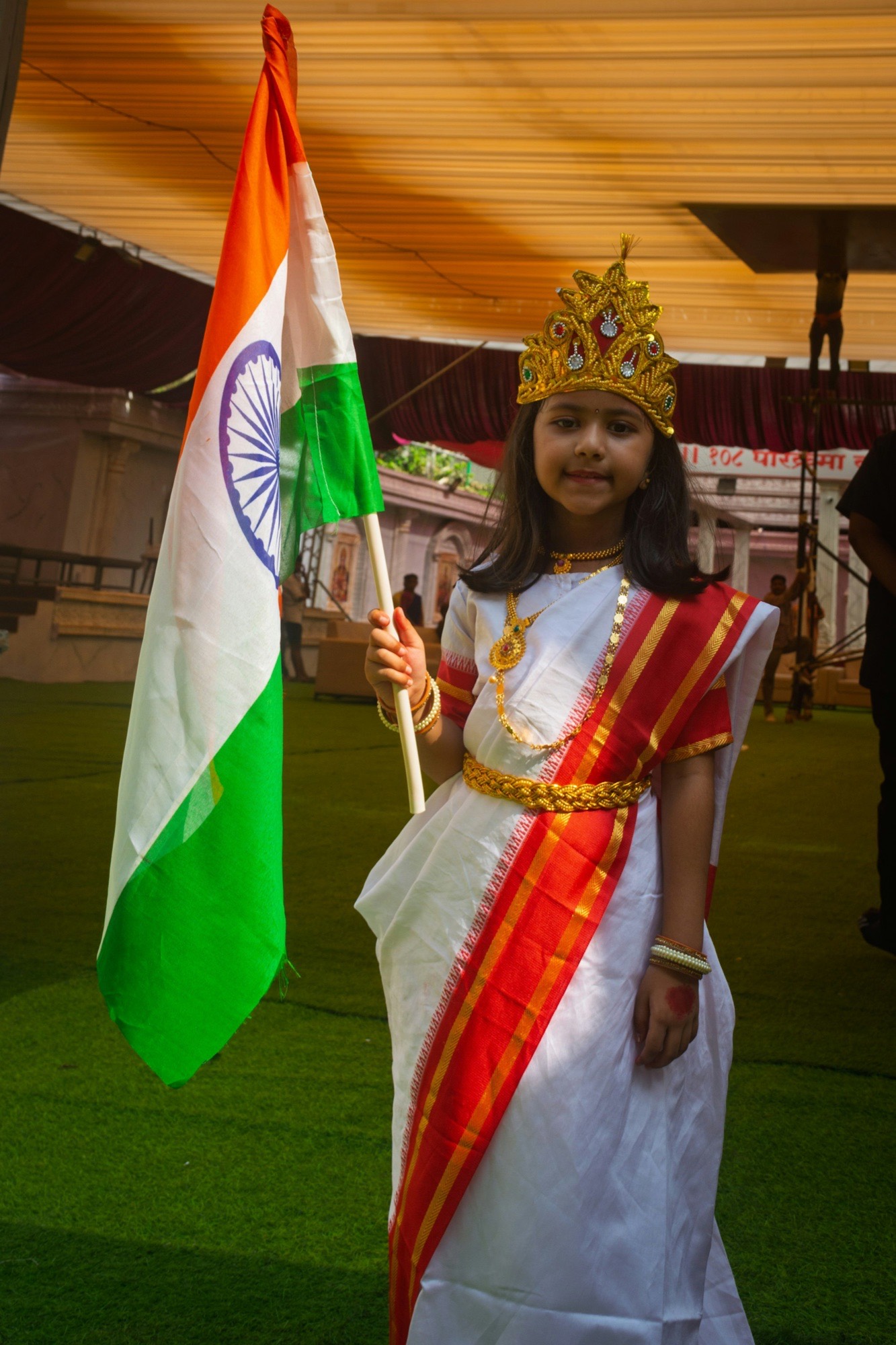 A young girl dressed as Bharat Mata in a white saree and gold crown, holding the Indian flag