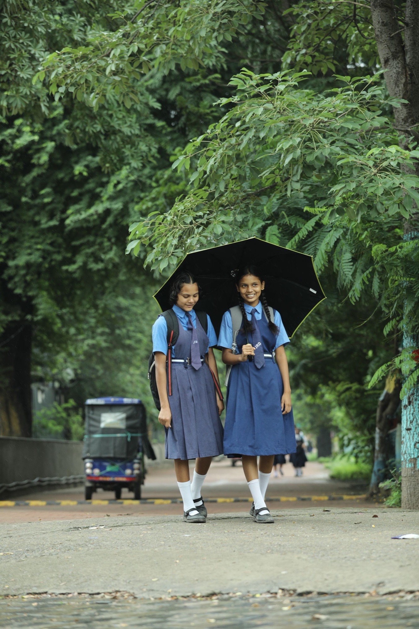 Two schoolgirls in blue pinafore uniforms sharing an umbrella on the way to school